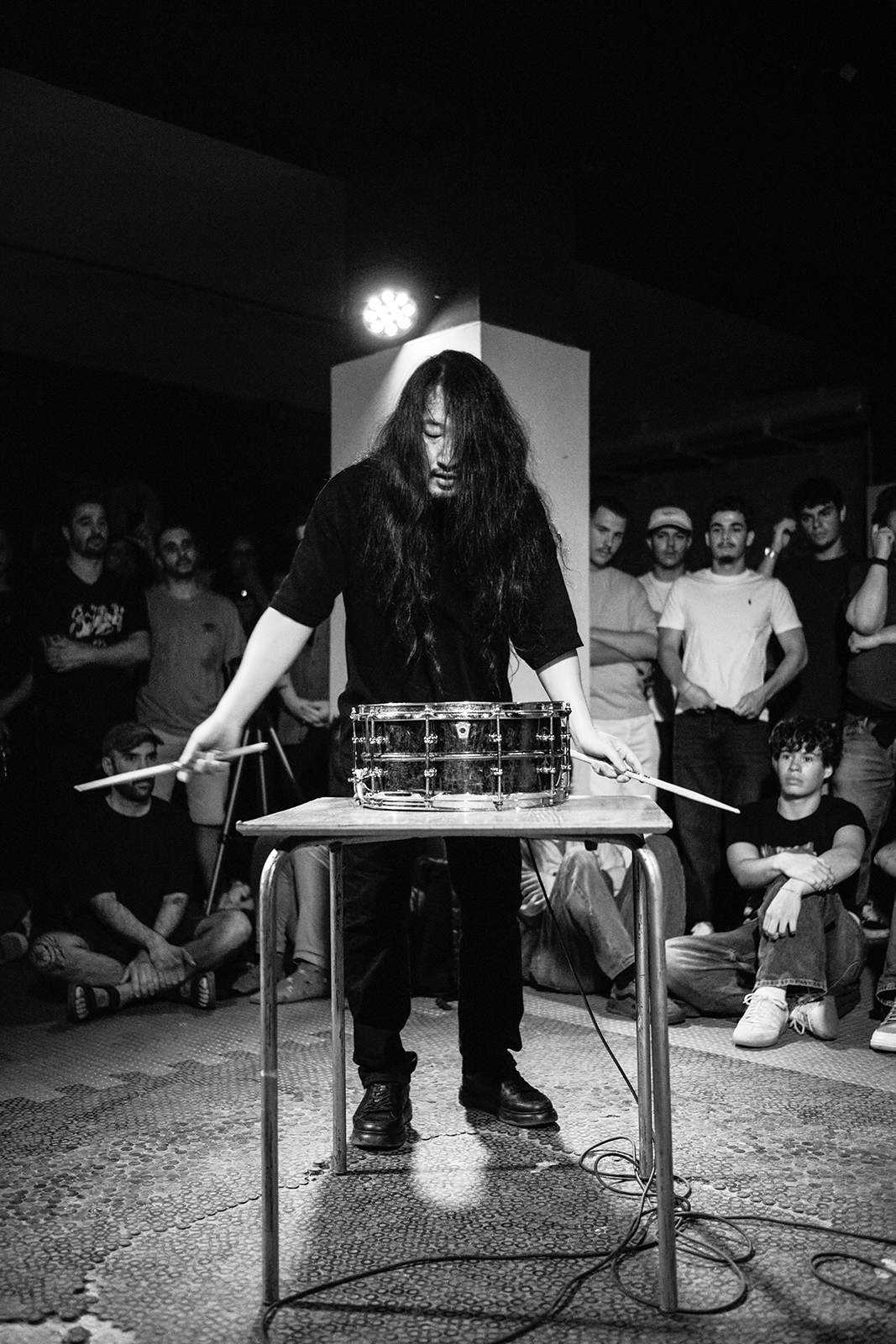 Músico com cabelo longo toca caixa de bateria sobre mesa, cercado por público em ambiente fechado.