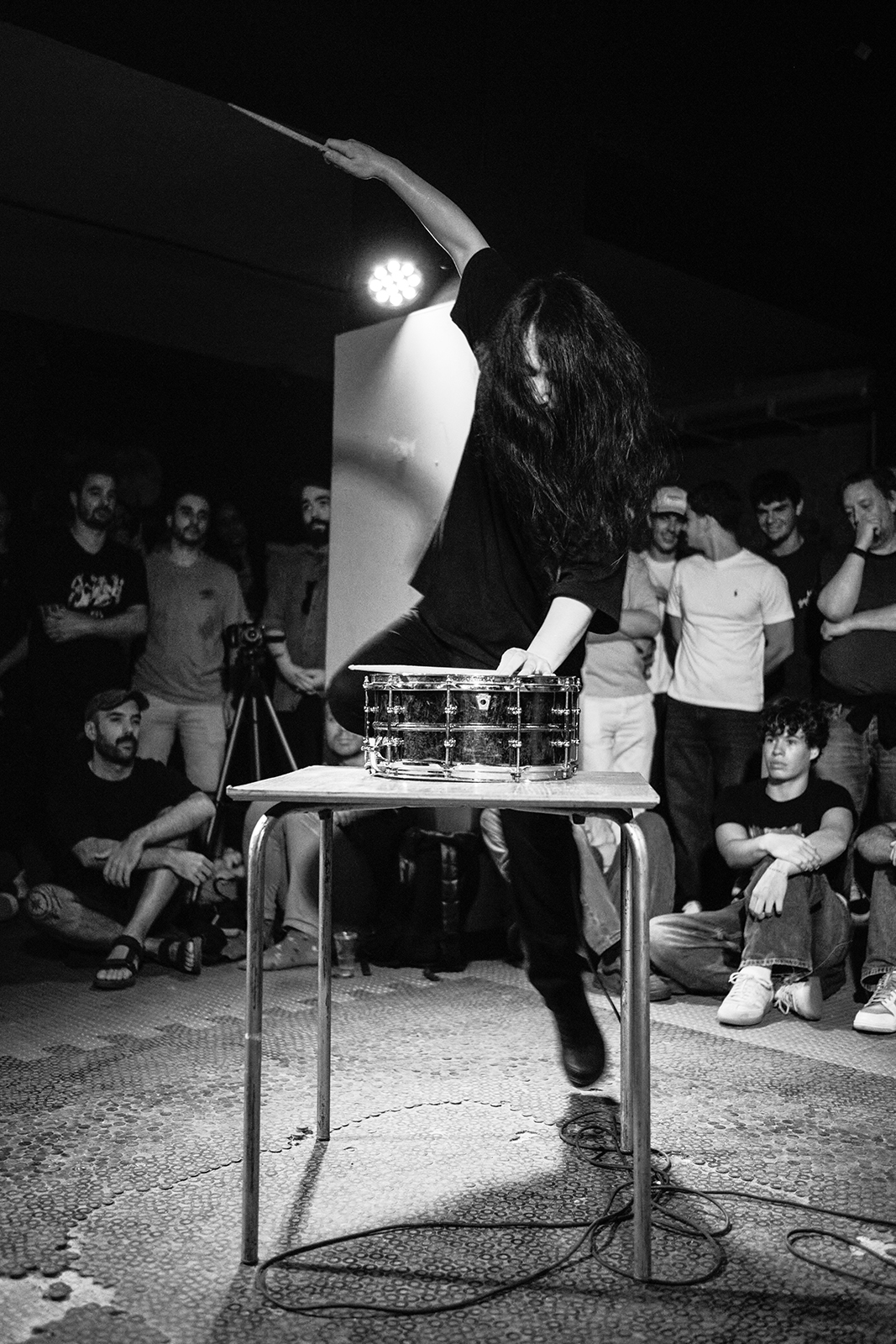 Pessoa toca caixa de bateria em ambiente escuro, cercada por espectadores atentos.