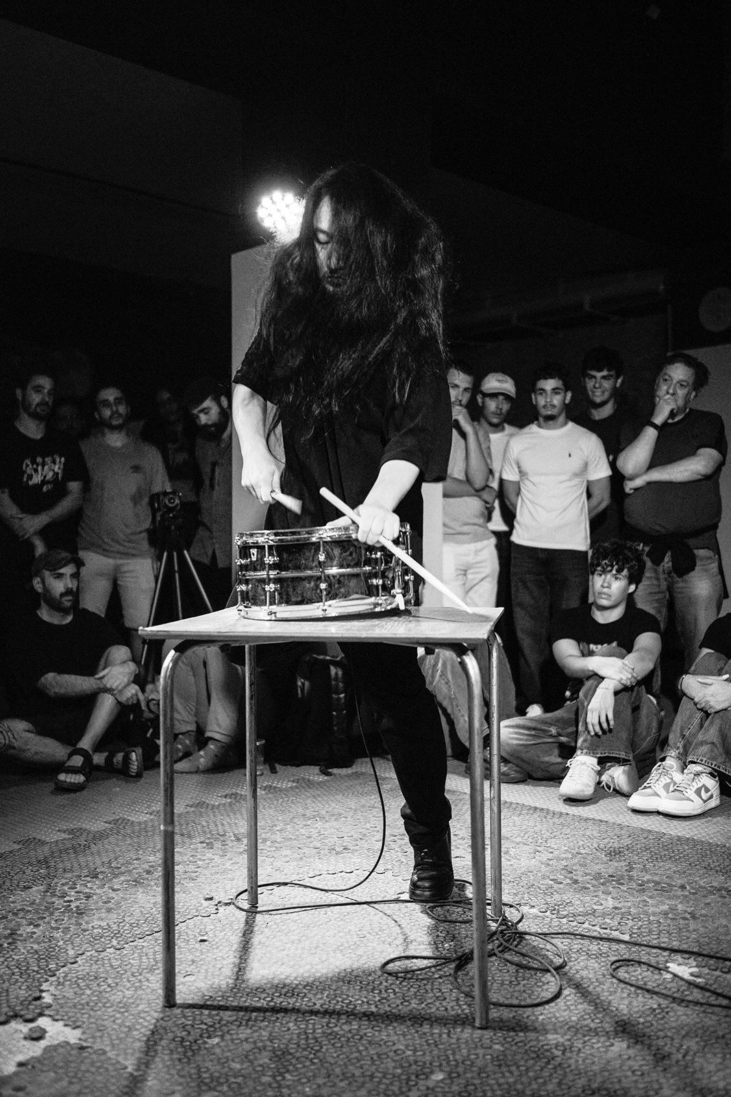 Músico toca tambor em mesa, público ao redor observando em preto e branco.