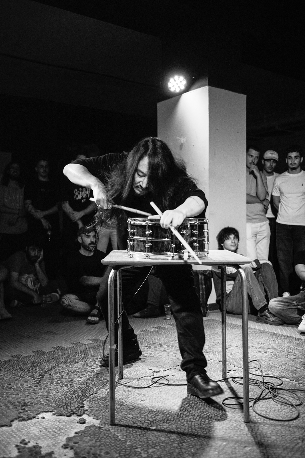 Músico toca bateria pequena sobre mesa com baquetas longas, cercado por espectadores.