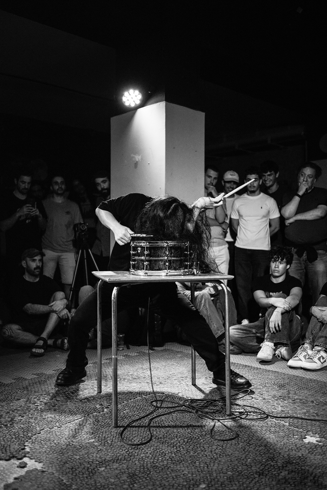 Artista toca tambor inclinado em performance ao vivo com plateia em ambiente escuro.