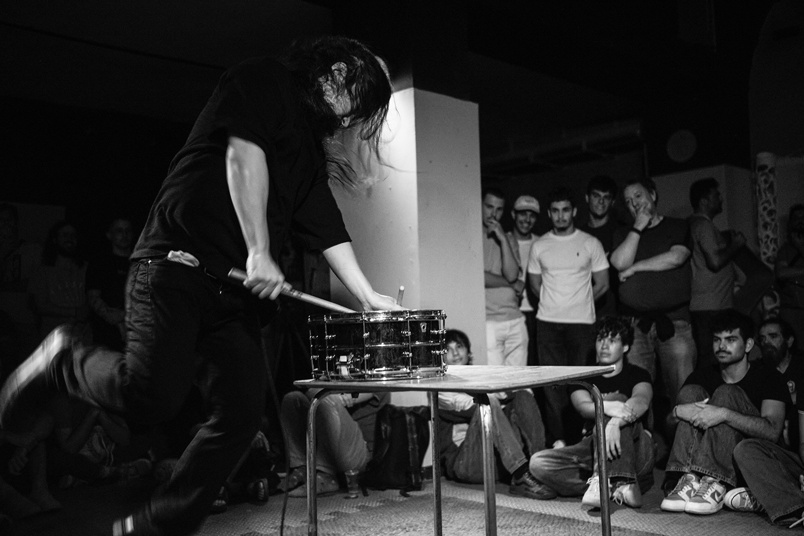 Músico tocando tambor com baquetas em show ao vivo, público observando em local fechado.
