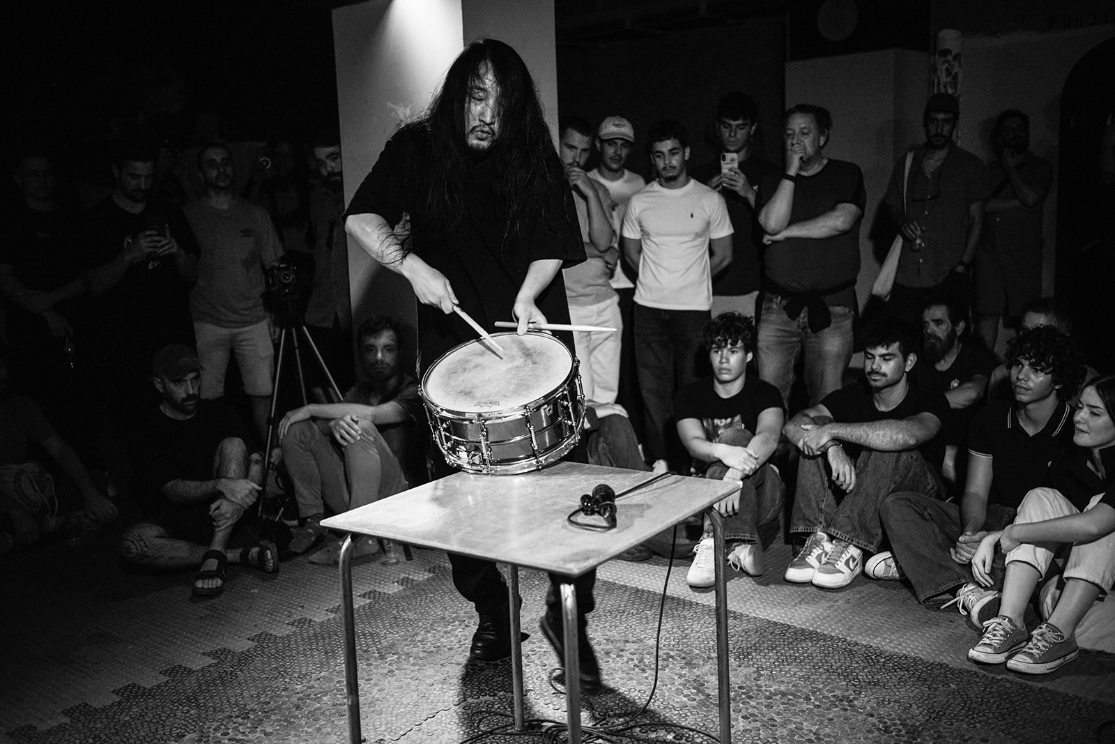 Homem com cabelo longo toca tarola em apresentação cercado por público atento em preto e branco.
