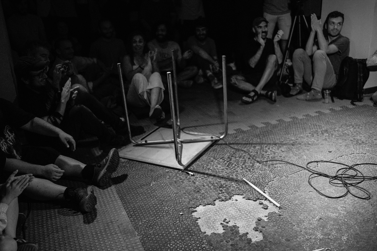 Pessoas sentadas aplaudem em sala, mesa virada e fios no chão, ambiente fechado.