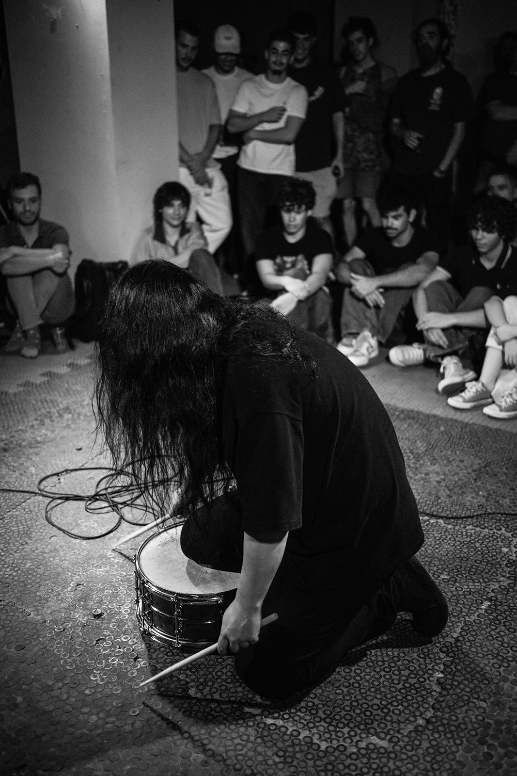 Pessoa ajoelhada tocando tambor com baquetas, grupo assiste em ambiente fechado.