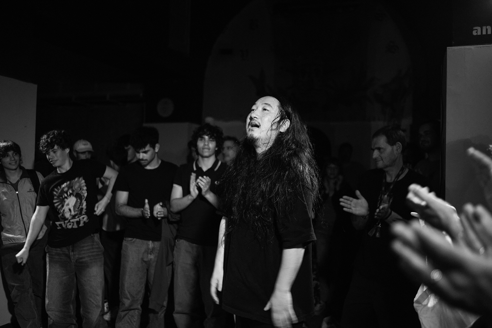 Pessoa de cabelos longos no centro de um evento sendo aplaudida, cena em preto e branco.